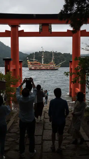 Tourists take photographs of a pirate ship cruise sailing on Ashinoko Lake through the Torii of Peace at Hakone Shrine.