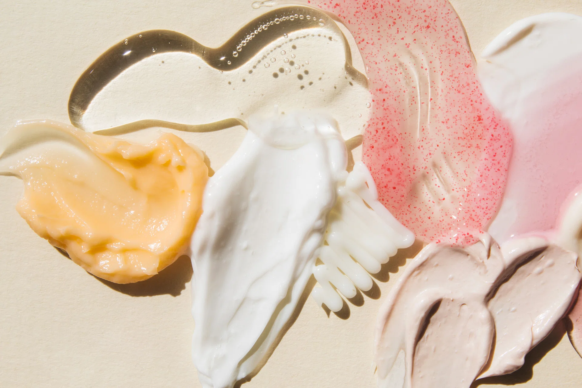 Textured multi colored smears of cosmetics on a beige background. Samples of creams, face mask, scrub with exfoliating particles, face gel, serum. Flat lay, top view.