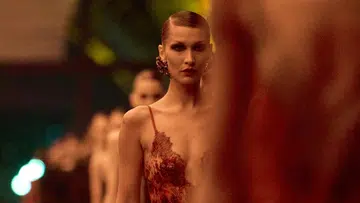 Bella Hadid walking the Saint Laurent Fall Winter 2026 runway in a sheer red lace lingerie-inspired gown with intricate floral embroidery and statement gold earrings.