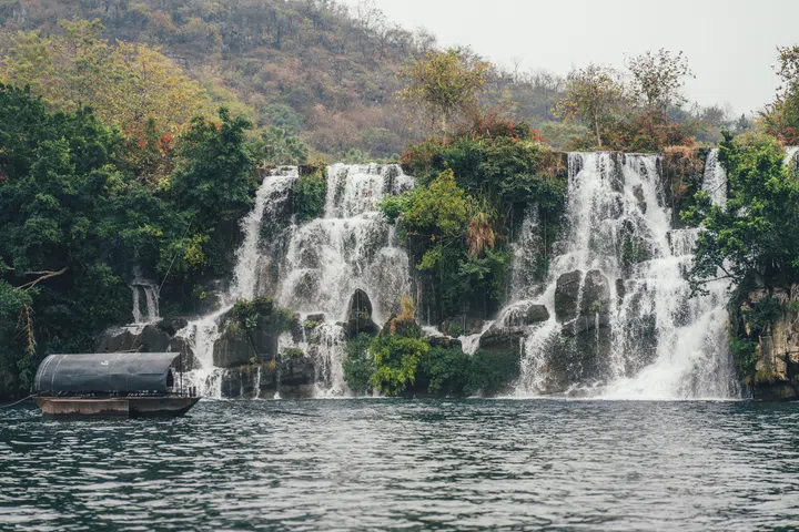 Liuzhou, Guangxi, China - January 30, 2025: This evocative image captures a vibrant, multi-tiered waterfall cascading into a tranquil lake in Liuzhou, Guangxi. Lush greenery, including dense foliage and flowering bushes with red blooms, frames the rushing water, which creates a misty spray against rugged, moss-covered rocks. A covered boat floats serenely on the calm, deep-green water, enhancing the peaceful atmosphere. The overcast sky casts soft, diffused light, highlighting the natural textures of the landscape from the smooth water s gentle ripples to the rough stone surfaces and blending earthy greens, whites, and grays with pops of red and yellow from the vegetation. The scene exudes tranquility, showcasing the region s rich biodiversity and serene natural beauty.