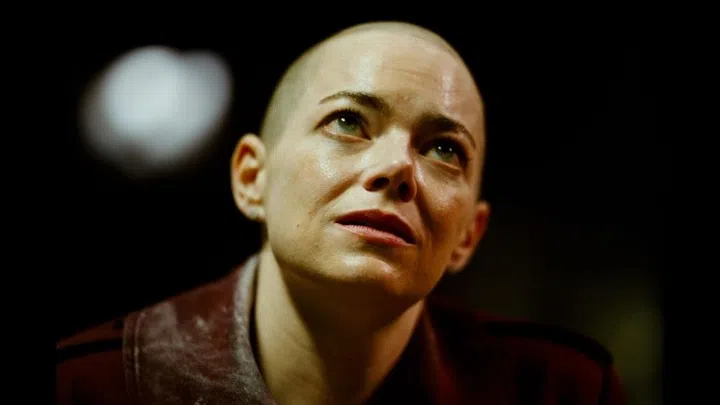 Emma Stone with a shaved head looking upward in a dramatic, close-up film still from Yorgos Lanthimos’s Bugonia.