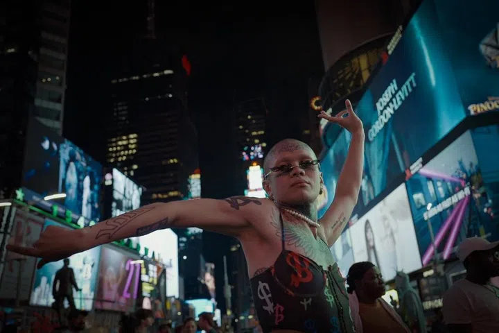 Documentary still from 10s Across the Borders showing a tattooed queer individual posing in Times Square. The image captures the intersection of identity and global urban spaces at night in New York City.