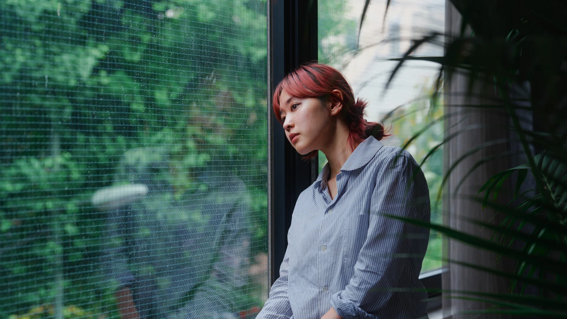 A sentimental portrait of a young woman standing next to a window. The weather outside is gloomy and overcast and rain, giving the photo sadness and nostalgia feeling.