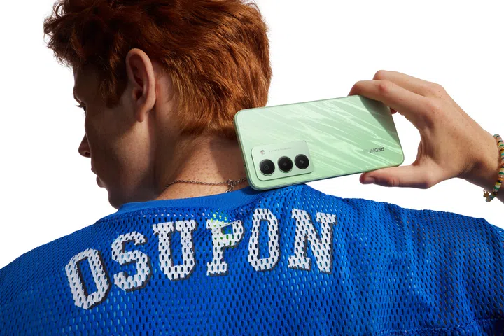 Model with back facing the viewer, holding a Ripple Green Redmi 15 5G phone and hovering the device over his right trapezius.