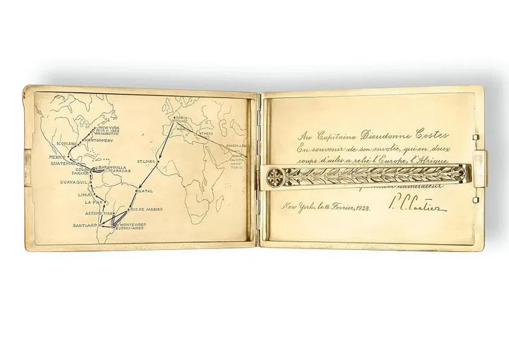 Cartier New York, circa 1928 — Gold and enamel - At the centre, the monogram DC, inside, an inscription and facsimile signature which translates as: "To Captain Dieudonné Costes / in memory of his flight, who with two flaps of his wings linked Europe, Africa / and America. / From his compatriot and admirer. / New York, February 16, 1928 / P. C. Cartier". It is accompanied by a map with blue enamel arrows indicating the stages of the path followed by the pilot. Photo: Cartier Collection