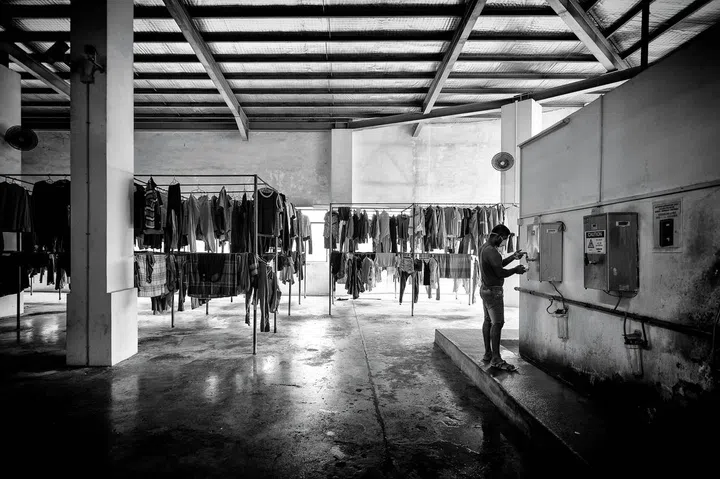 A migrant worker at a hot water dispenser amidst drying clothes at a Factory-Converted Dormitory. Residents are encouraged to wash both themselves and their clothing often to maintain hygiene levels and laundry detergent and body soap are some of the most requested items from NGOs and volunteer groups doing distribution. Photo: Bryan van der Beek
