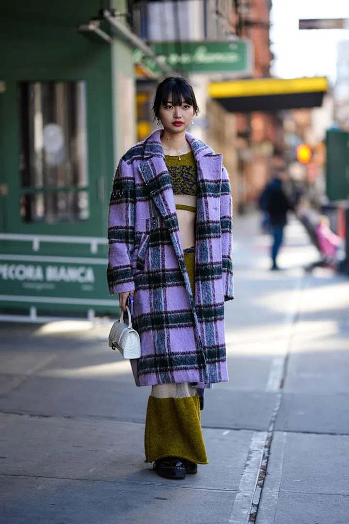 NEW YORK, NEW YORK - FEBRUARY 11: A guest wears gold pendant earrings from Chanel, a khaki with embroidered navy blue pattern cut-out / long sleeves / cropped top, a matching wool khaki with embroidered navy blue pattern long tube skirt, a white shiny leather crocodile print pattern Hourglass handbag from Balenciaga, black shiny leather varnished laces ankle boots from Dr Martens, a pale purple and green / brown checkered print pattern wool oversized long coat , outside PH5, during New York Fashion Week, on February 11, 2023 in New York City. (Photo by Edward Berthelot/Getty Images)