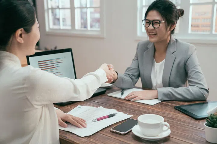 Two happy asian businesswomen shaking hands in cafe bar by window. office ladies team workers sitting at table in modern coffee made good deal agreement with laptop computer chart. smiling female.