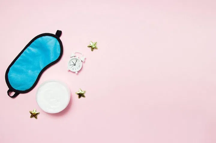 Blue sleeping eye mask, alarm clock and jar of cream on pink background. Night skin care concept.