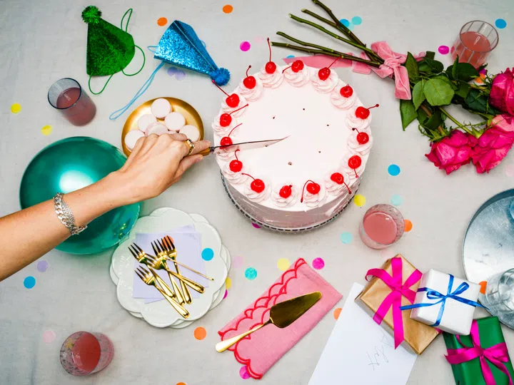 High Angle View Woman's Hand Slicing Into Pink Cherry Birthday Cake On Table With Roses And Gifts At Party