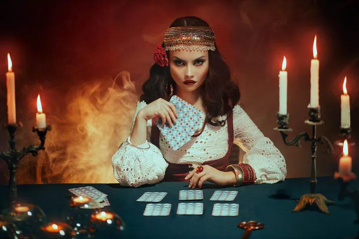 Fantasy beautiful magnificent girl in image of a gypsy sits at table in dark gothic room. Red costume, vintage clothing, art black make up. Fortune teller woman reading future on magical tarot cards
