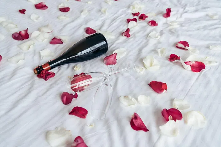 rose petals, champagne glass and bottle of champagne on white sheet on bed in bedroom