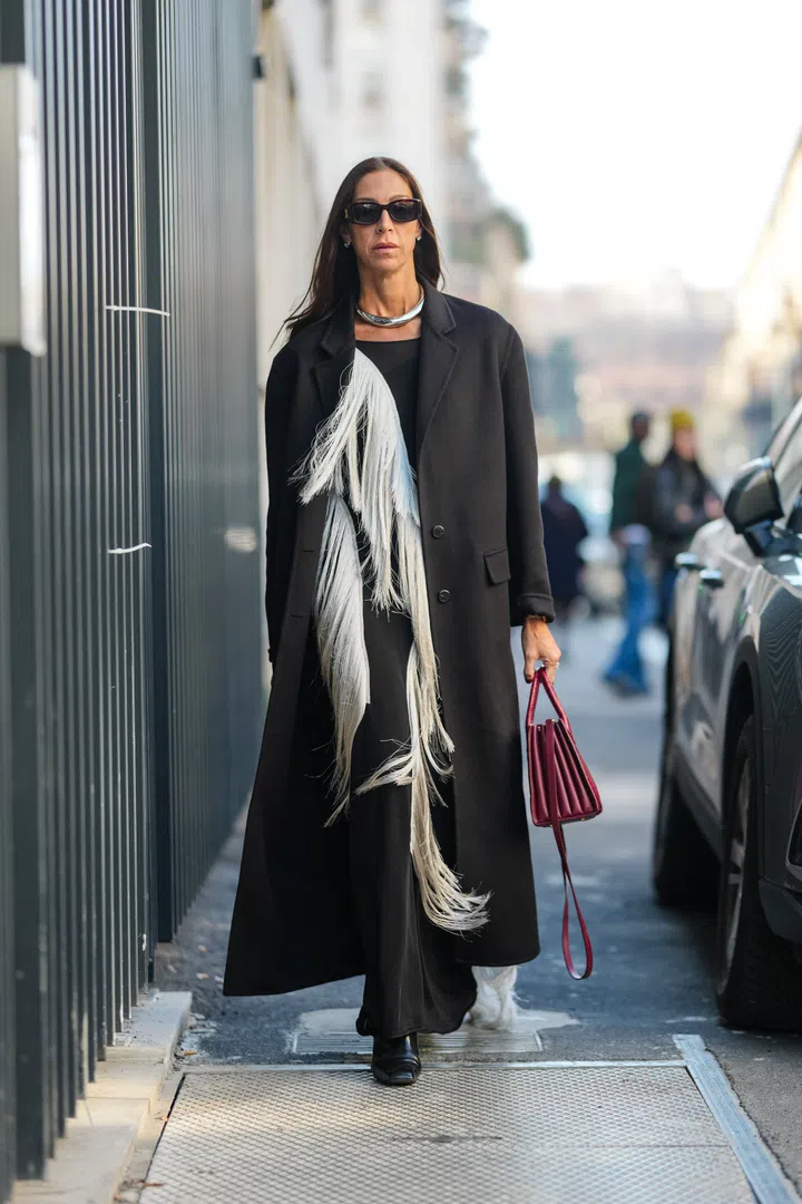 MILAN, ITALY - FEBRUARY 27: A guest wears black sunglasses, white gold diamond earrings, silver metallic necklace, black oversized wool coat, black with white fringe midi dress, shiny burgundy bordeaux leather bag, outside Blumarine, during the Milan Fashion week Women's Fall/Winter 2025-2026 on February 27, 2025 in Milan, Italy. (Photo by Edward Berthelot/Getty Images)