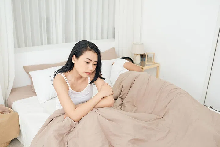 We have a problem. Young upset girl sitting on the edge of the bed, against her boyfriend, lying in bed.