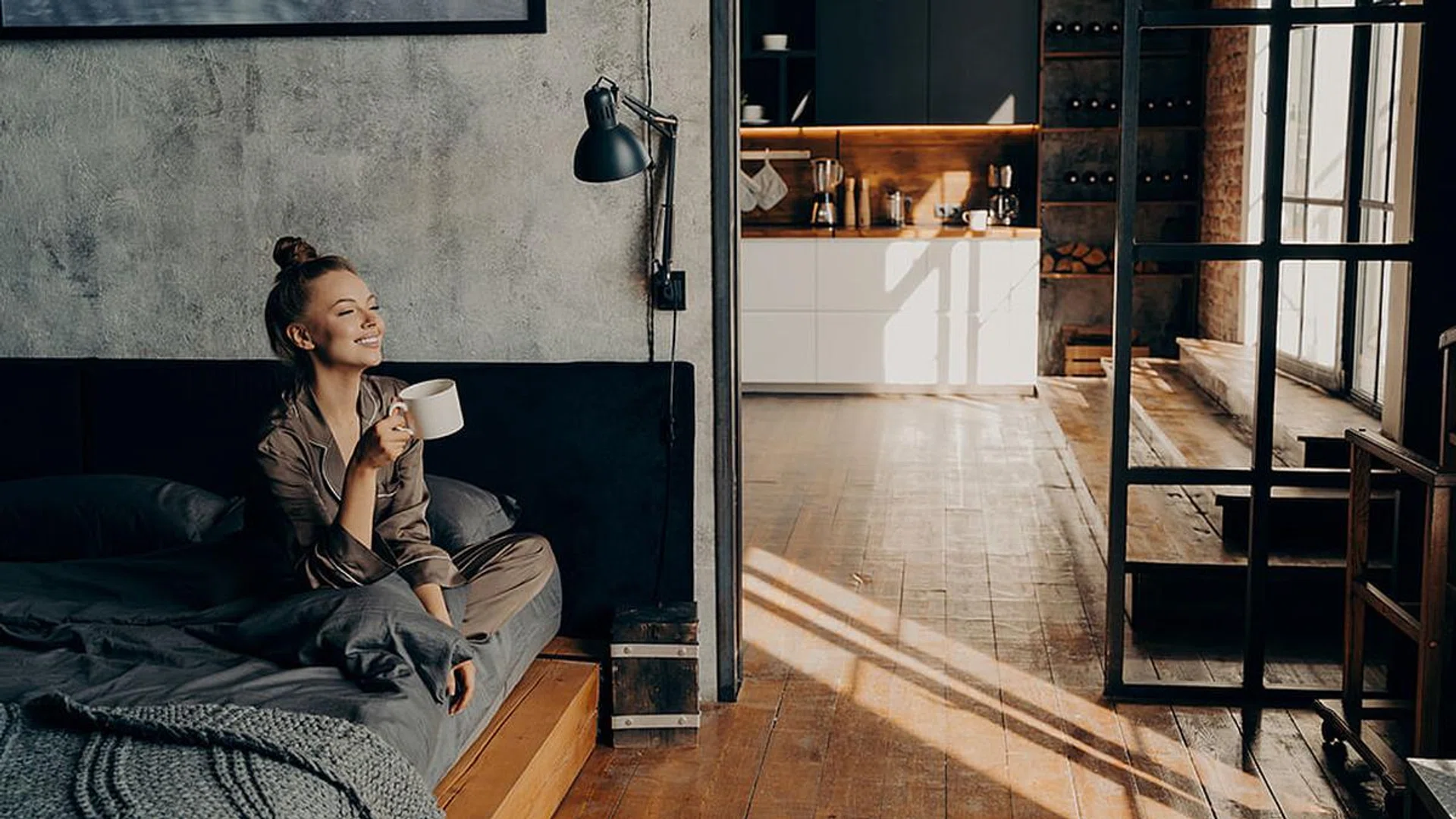 Young happy attractive girl sitting on bed in satin pajama in lotus position with cup of hot coffee in her hand while looking toward window smiling at morning sunlight, enjoying weekend at home