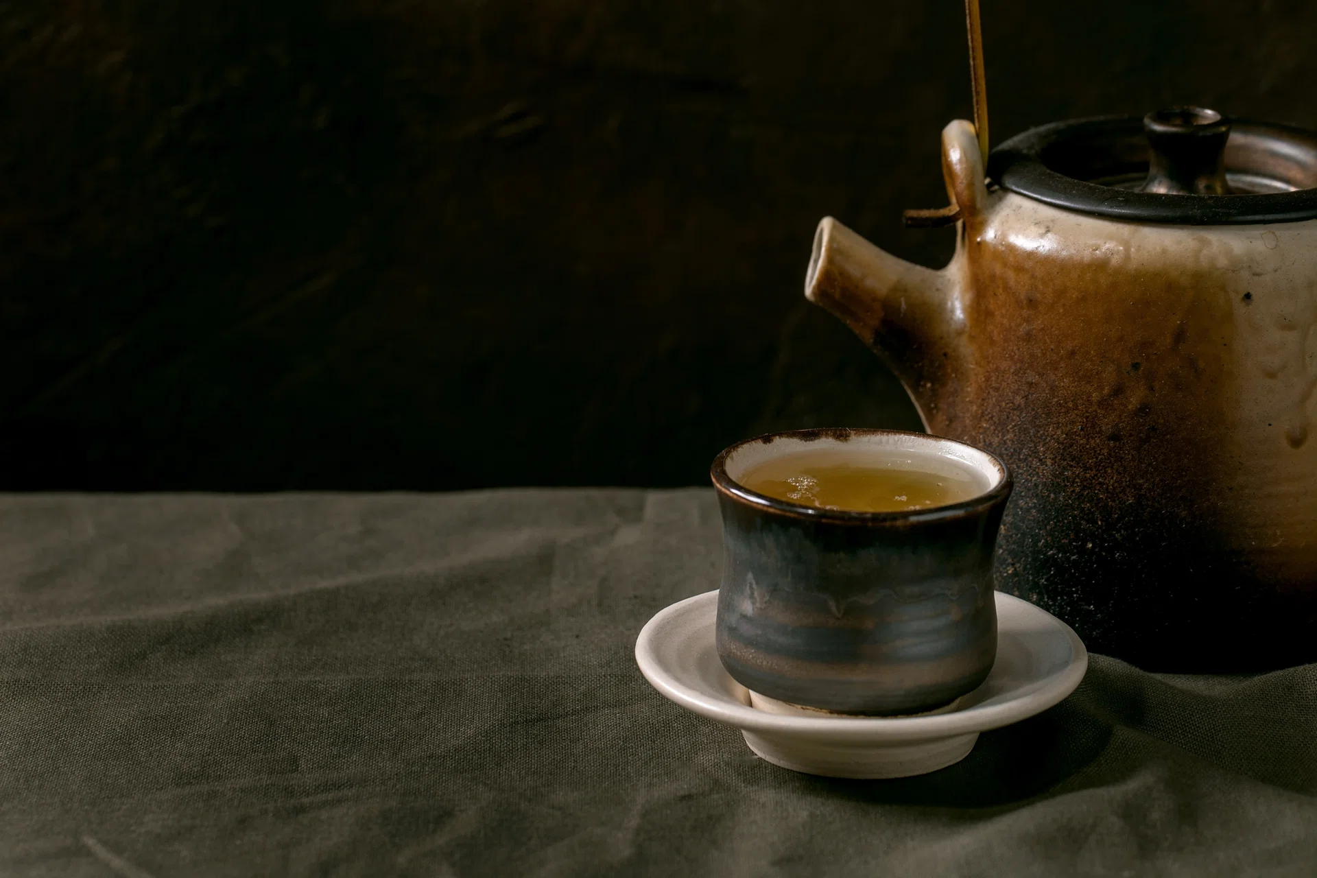 Craft handmade ceramic teapot kettle with cup of hot green tea with saucer, standing on linen tablecloth in dark room, Close up. (Photo by: Natasha Breen/REDA/Universal Images Group via Getty Images)