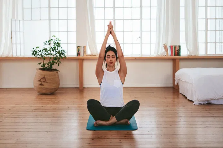 Full length shot of healthy and beautiful young woman sitting at home in yoga position. Fitness female sitting with hands joined above her head.