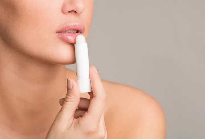 Woman applying hygienic balm to her plump lips on grey studio background, free space