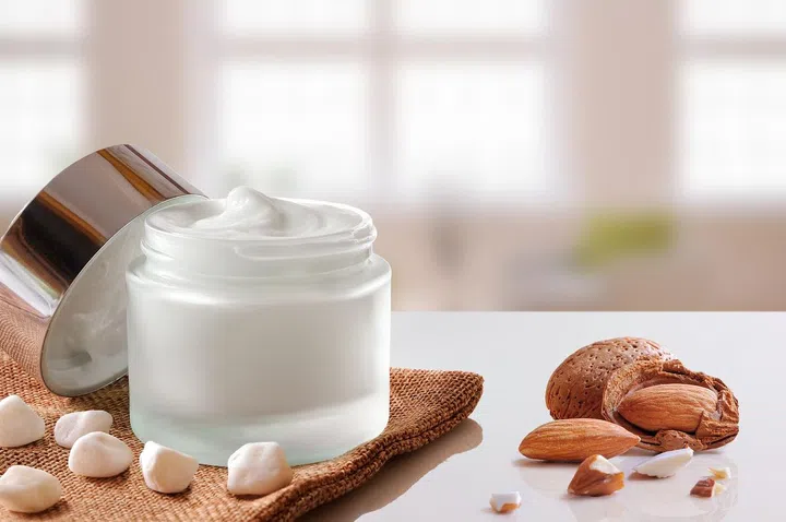 Glass open jar with facial or body almond moisturizer on burlap. With lid, small stones and almonds. Windows background. Front view.