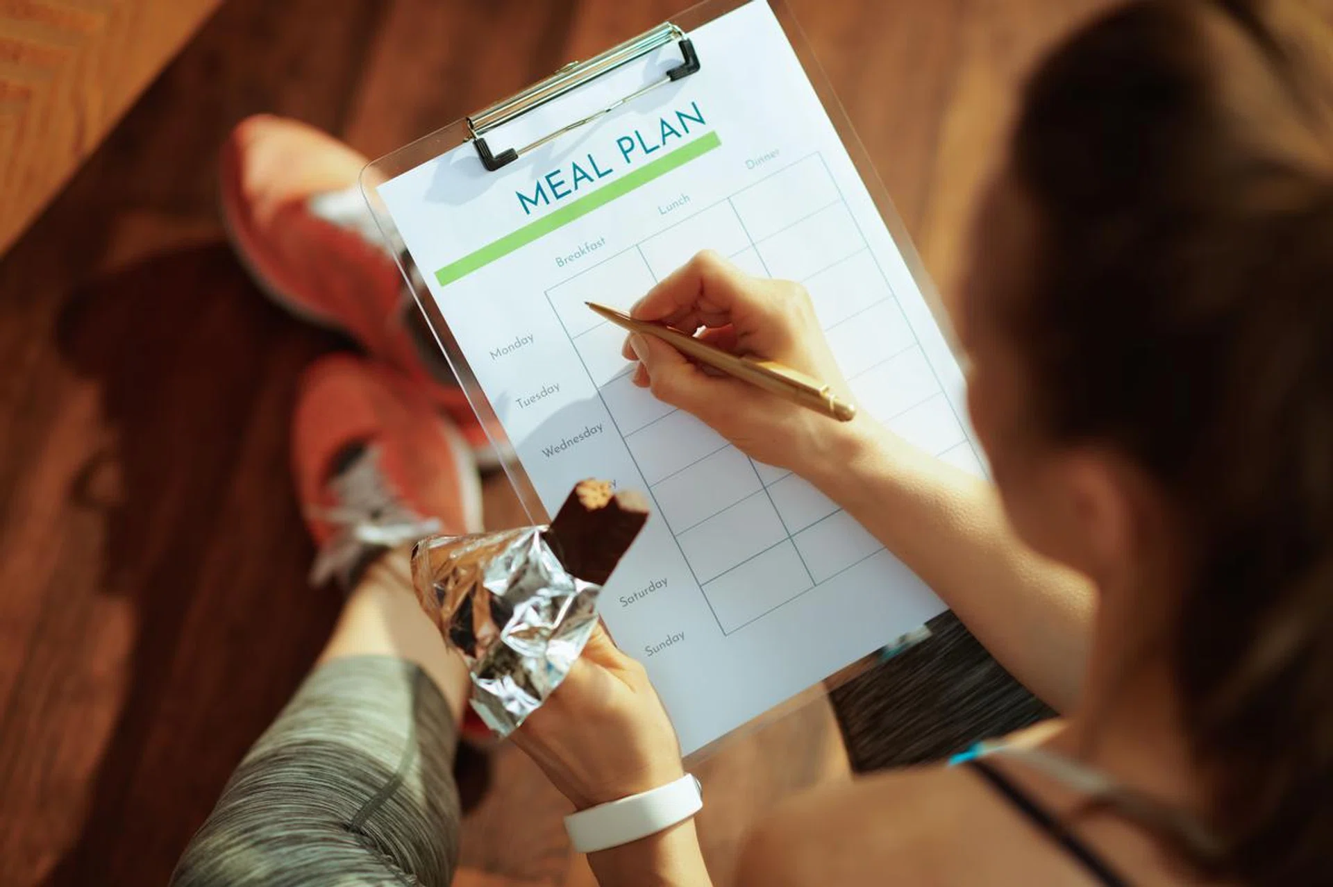 Seen from behind fit sports woman in sport clothes with chocolate raw protein bar filling meal plan in the modern living room.