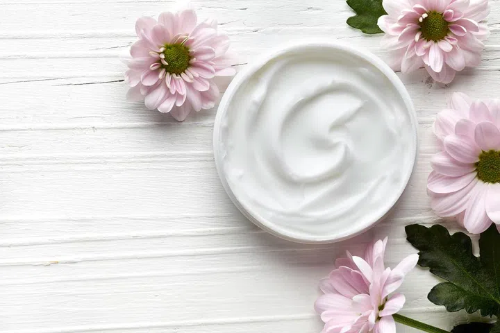Cosmetic cream container and pink flowers on white wooden background from top view