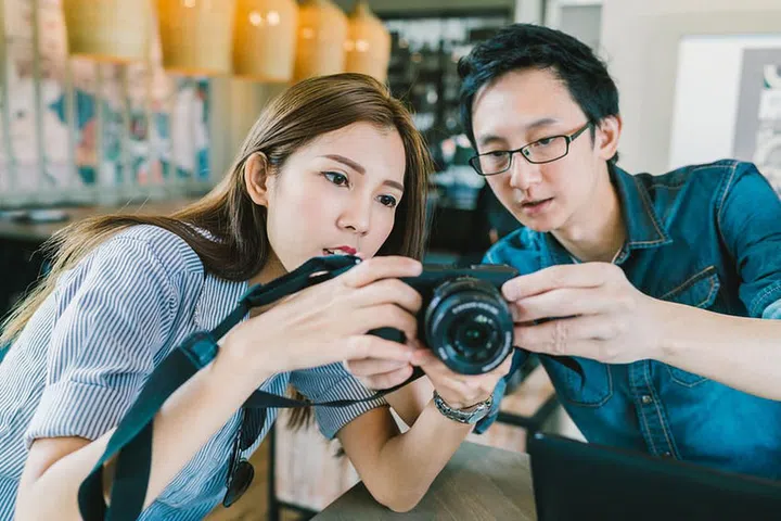 Young Asian couple learning to use mirrorless digital camera together at coffee shop, modern gadget technology concept, focus on the girl, depth of field effect