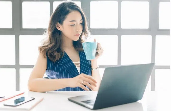 Portrait of beautiful Asian businesswoman sitting near bright window while looking at open laptop computer on table and with cup of coffee.