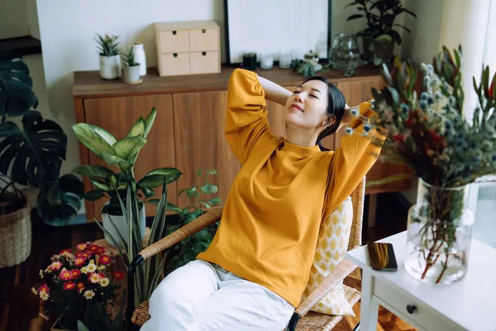 Young Asian woman with hands behind her head, taking a break and relaxing on armchair at cozy home, surrounded by green houseplants. Lifestyle, wellbeing and wellness concept