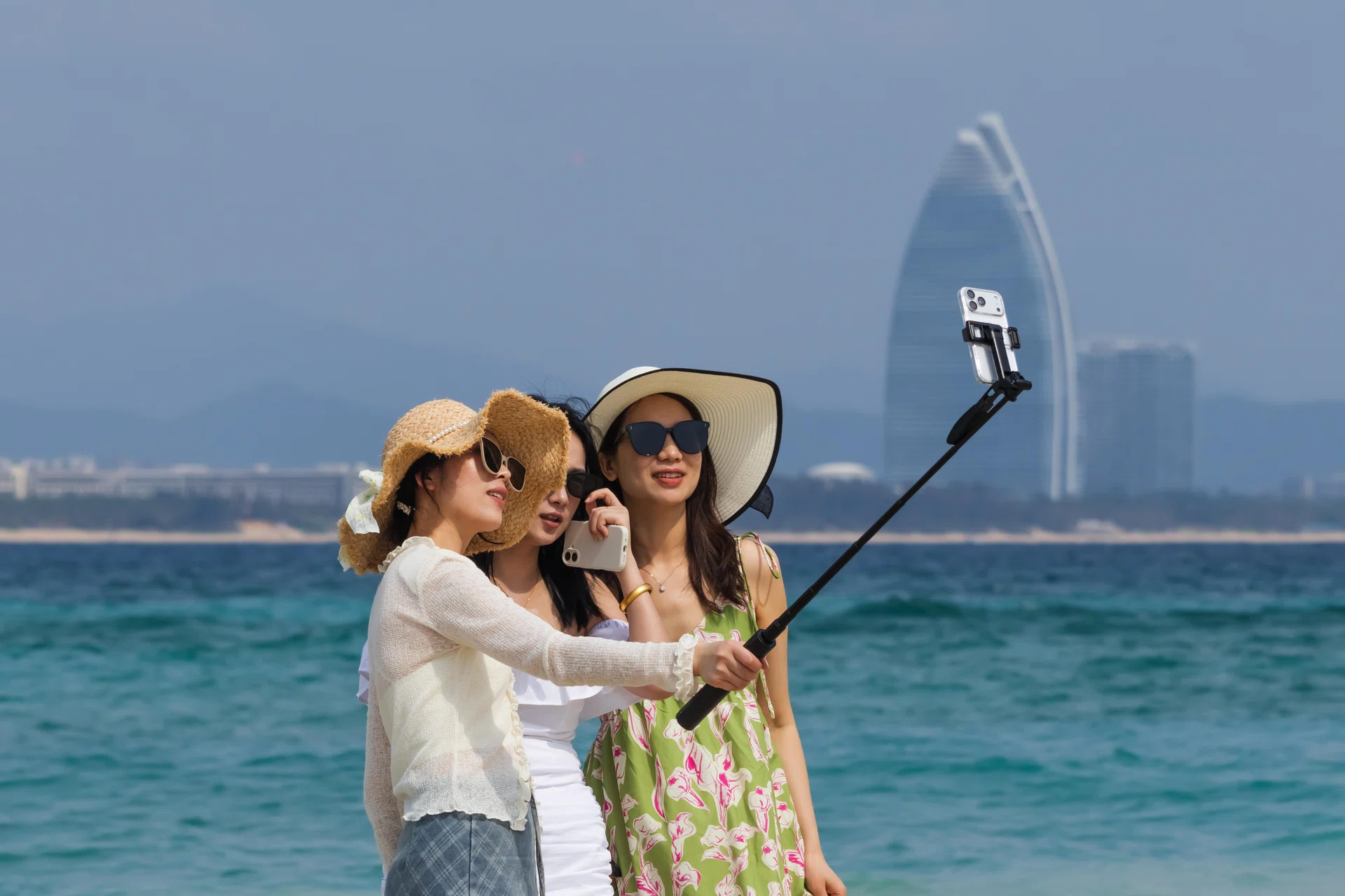 SANYA, CHINA - MARCH 7: Three young women wearing sun hats and sunglasses take a selfie with a smartphone mounted on a selfie stick while standing on the beach at Wuzhizhou Island, with the Atlantis Sanya resort visible in the background across the sea, on March 7, 2026 in Sanya, Hainan Province, China. Wuzhizhou Island is a popular tourist destination known for its tropical scenery and marine tourism activities along the South China Sea. (Photo by Cheng Xin/Getty Images)