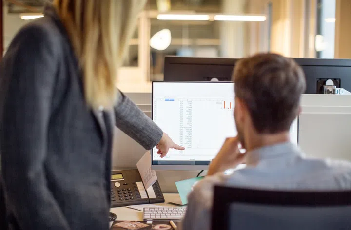 Rear view of two business people working together on computer. Woman pointing at computer monitor with man sitting by in office.