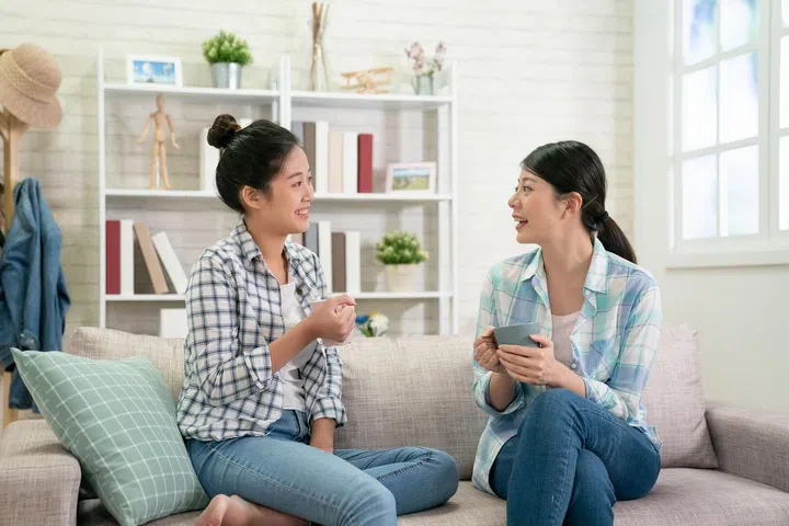 two happy young asian female friends with coffee cups conversing in living room at home. group of beautiful girls bestie sitting on couch relax in bright cozy apartment enjoy tea in weekends gossip