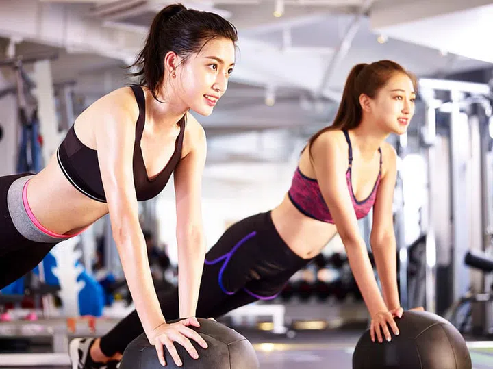 two young asian adult women working out in gym using medicine balls.
