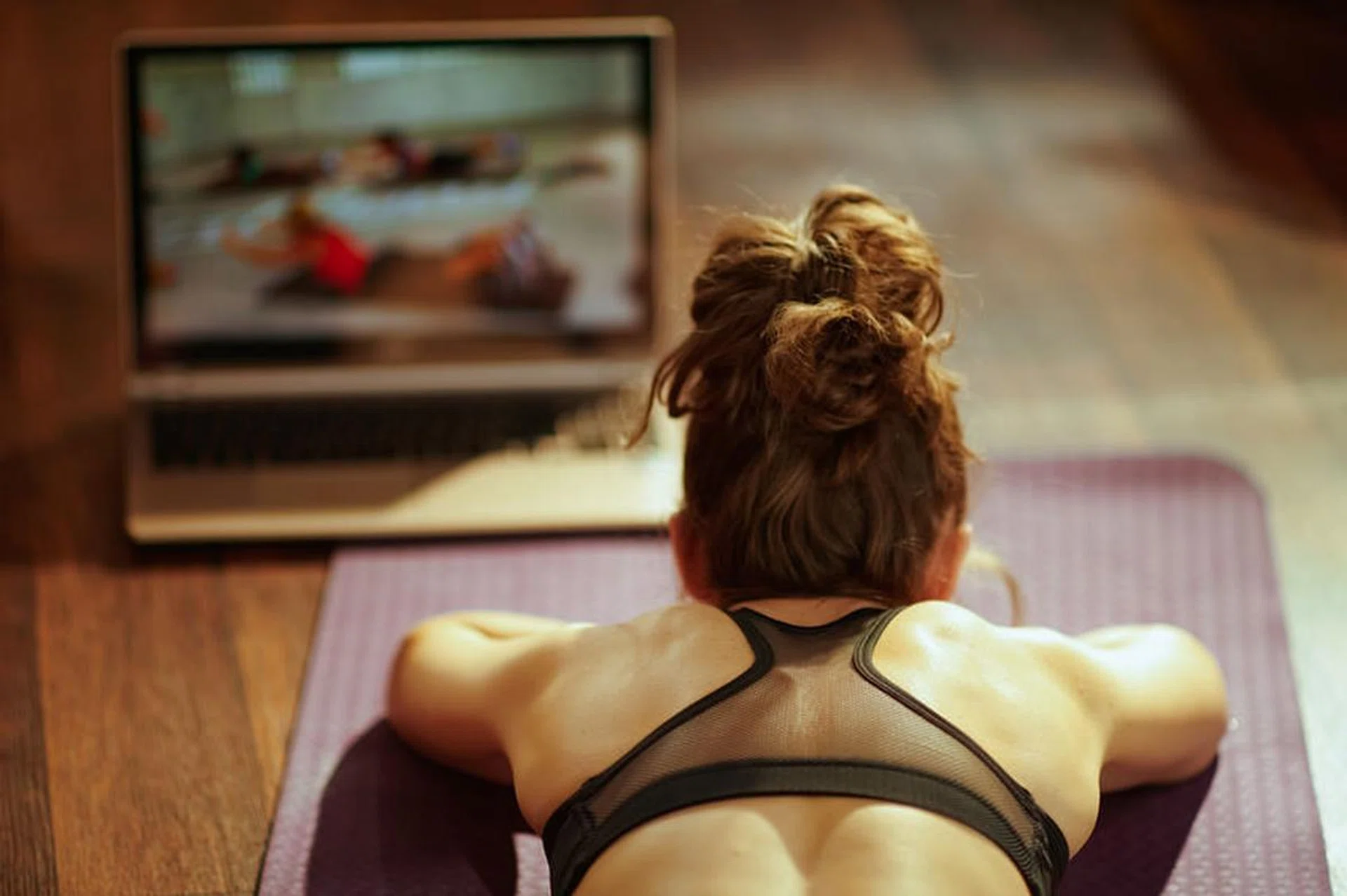 Seen from behind fit sports woman in fitness clothes in the modern living room using online fitness training program in laptop while laying on fitness mat.
