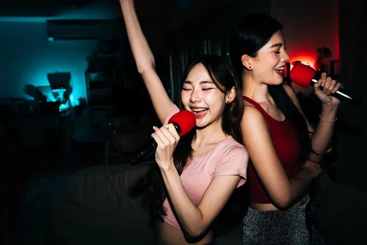 Joyful young women singing karaoke with red microphones in flash-lit room