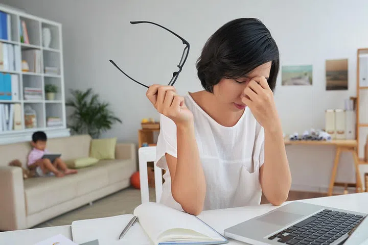 Stressed tired woman at her workplace, her child sitting on sofa behind her