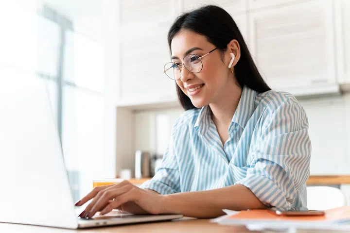 Image of a beautiful happy brunette young woman at the kitchen indoors at home using laptop computer and wireless earphones.