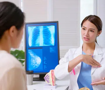 attractive young brunette ponytail female asian doctor wear white coat with pink ribbon explaining breast self examination to woman at clinic - a model on table and xray display