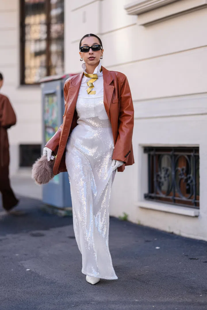 MILAN, ITALY - FEBRUARY 27: A guest wears black sunglasses, silver earrings, white buttoned up long sleeve shirt, gold metallic necklace, shiny dark brown leather blazer jacket, shiny white sheer mesh sequin midi dress, white tights, white pointed toe heels leather shoes, light brown faux fur bag, white leather gloves, outside Max Mara, during the Milan Fashion week Women's Fall/Winter 2025-2026 on February 27, 2025 in Milan, Italy. (Photo by Edward Berthelot/Getty Images)