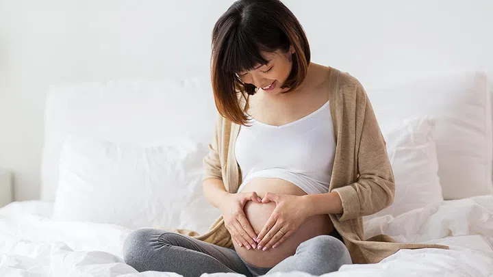 pregnancy, love, people and expectation concept - happy pregnant asian woman sitting in bed and making hand heart gesture at home