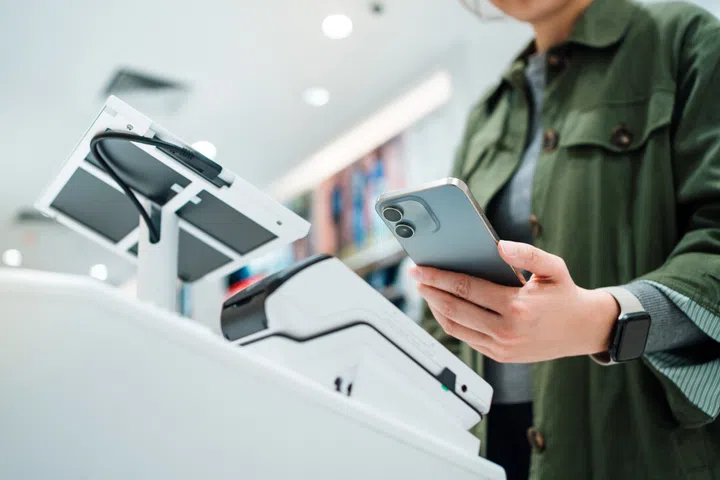 Close up of a woman's hand making mobile payment with her smartphone in a shop, scan and pay a bill on a card machine making a quick and easy contactless payment at self checkout counter. NFC technology, tap and go concept