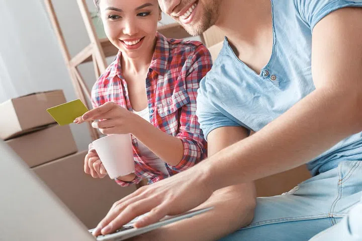 Young husband and wife moving to new place sitting on floor browsing laptop shopping online holding debit card drinking hot coffee smiling cheerful close-up