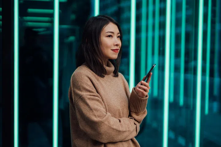 Young woman using mobile phone against futuristic holographic background. New ways of investing - using mobile app to buy stock and cryptocurrency.
