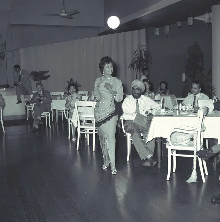The sarong need not always be made from batik as Vicki Dutton demonstrates. Here she wears an outfit especially chosen for the modern Malayan miss. Cut from cool, crush-resistant linen, the kebaya is snuggly fitted with collarless neckline and the knee in front. Over one hip is draped an apron-like panel, edged with a wool fringe. The apron is embroidered with black and white wool flowers, as are the skirt slit, neckline and cuffs. For extra glamour, there is a wide black chiffon bow at the waist.