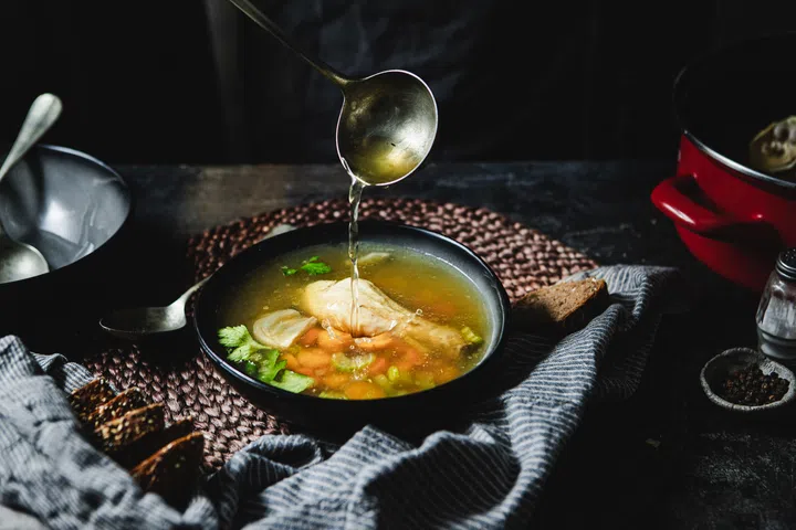 Cropped shot of pouring soup in bowl using ladle. Serving chicken broth in soup bowl over kitchen counter.