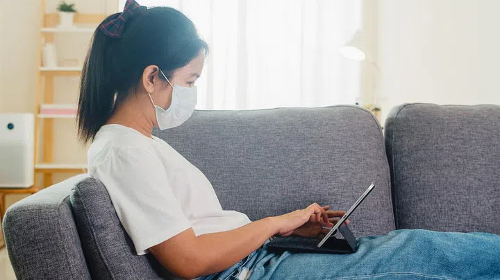 Asian freelance business woman wearing protective mask using tablet working in living room from home when social distancing stay at home and self quarantine time, coronavirus concept.