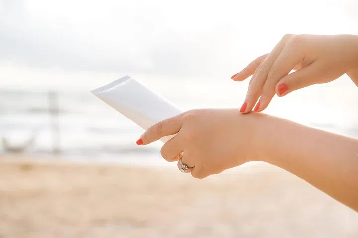 Woman hand holding sunscreen on the beach with the sea in blue sky background, spf sunblock protection and skin care concept
