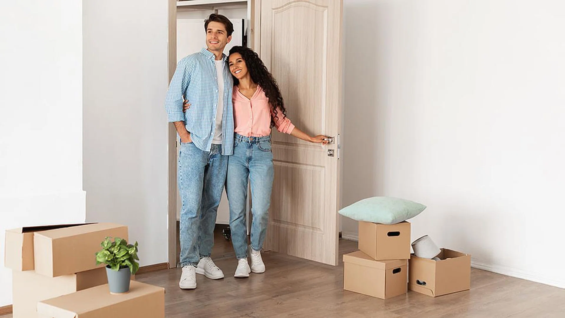 Housing Concept. Happy couple walking in empty house with cardboard carton boxes on floor, young family moving into new apartment after buying house, excited guy and lady hugging, looking around flat