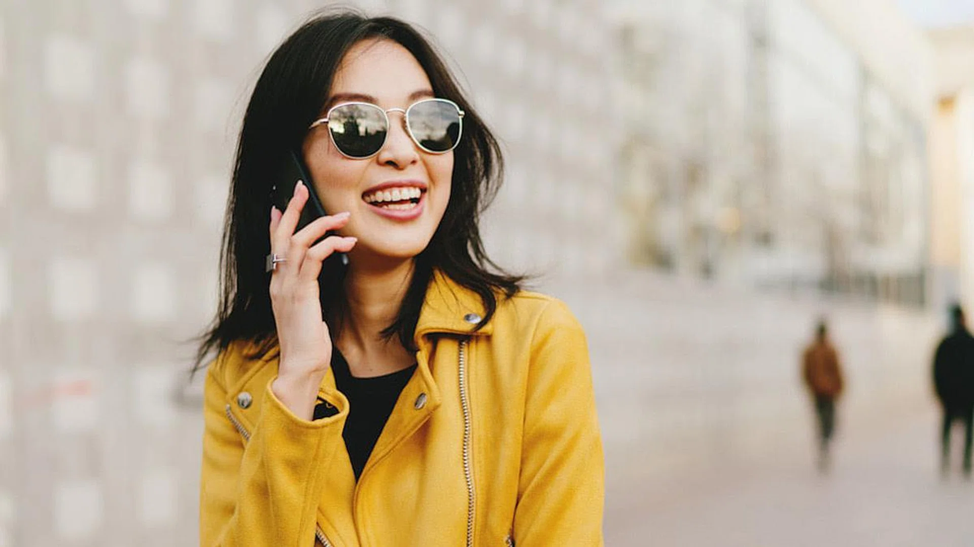Smiling asian woman with long dark hair in sunglasses talking by a smartphone while walking the city street. Young entrepreneur making business calls by a mobile phone while going to coworking space.