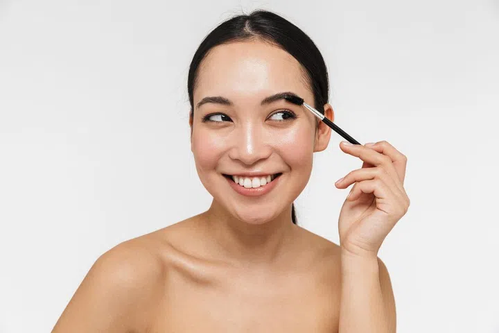 Photo of a beautiful young pretty asian woman with healthy skin posing naked isolated over white wall background holding brow styler.
