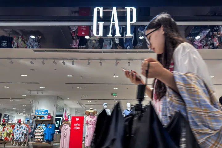 CHONGQING, CHINA - MAY 31: A woman looks at her phone while walking past a GAP retail store with mannequins and colorful clothing displays visible inside, on May 31, 2025 in Chongqing, China. GAP Inc. recently reported weaker-than-expected quarterly earnings and announced further store closures globally as it shifts focus toward e-commerce and streamlining operations to combat slowing consumer demand and rising operational costs. (Photo by Cheng Xin/Getty Images)